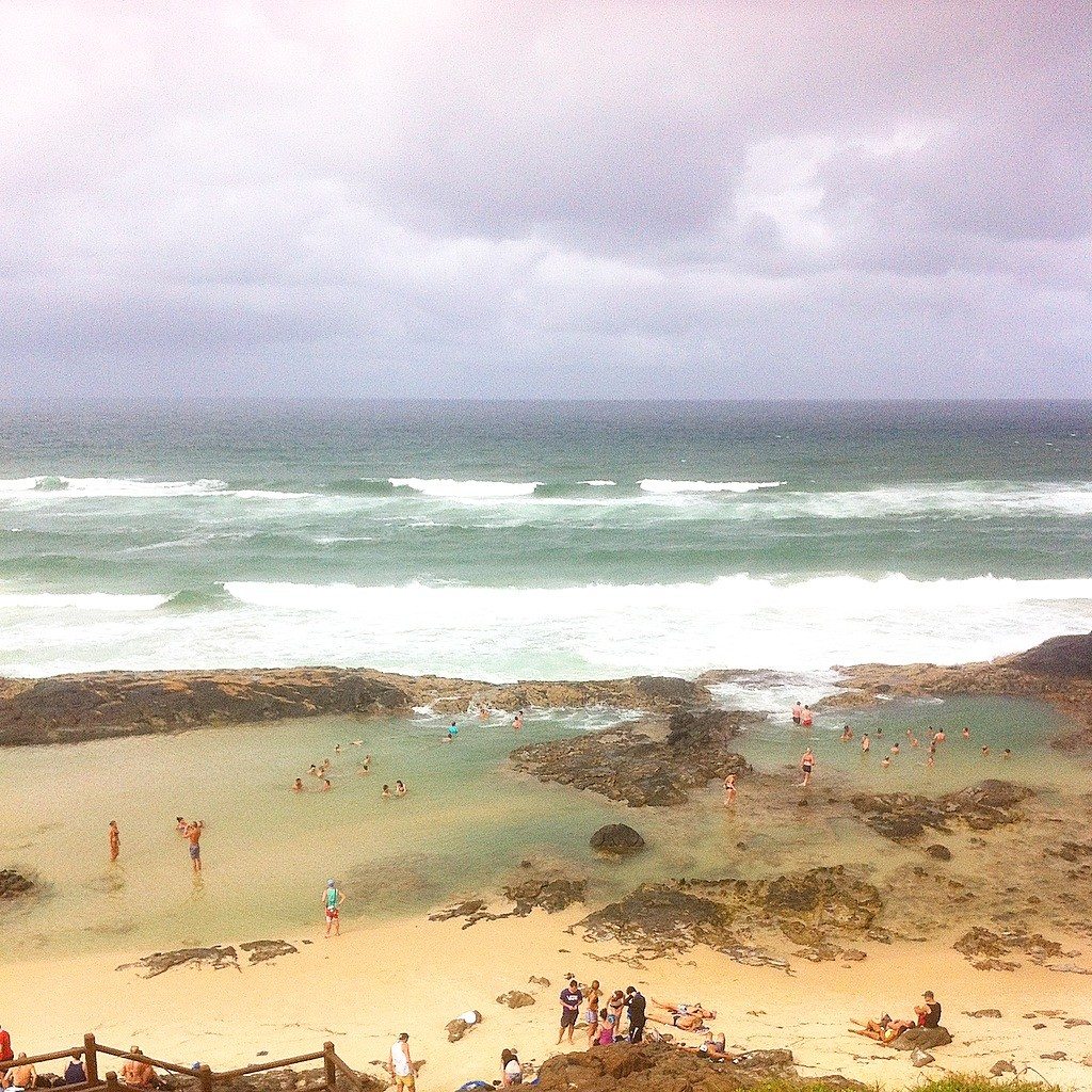 Pessoas nadando e relaxando em uma piscina de pedra na praia em Fraser Island, com ondas no oceano e céu nublado.