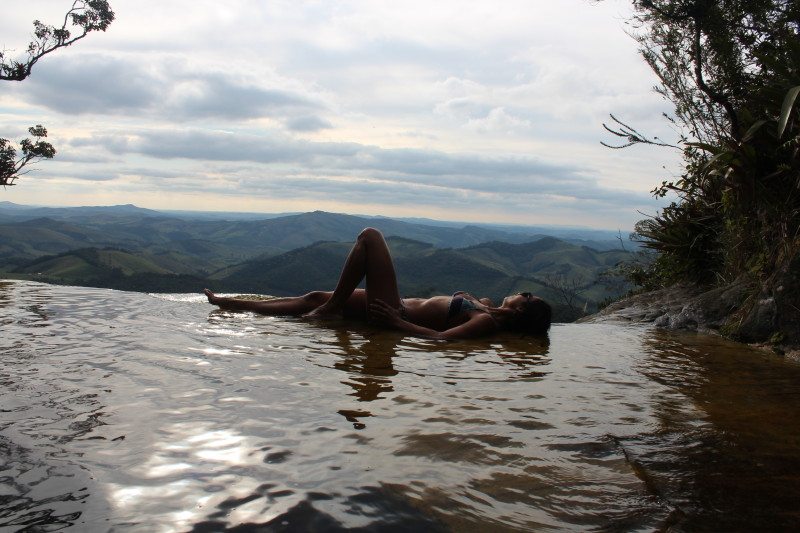 Atração do Parque Estadual do Ibitipoca que chama Janela do céu onde uma mulher está deitada na água com uma borda infinita. A vista da cachoeira dá para as montanhas e para o céu limpo durante o dia. Imagem ilustrando post O que fazer em Ibitipoca.