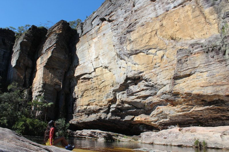 Paredão de pedra dentro do Parque Estadual do Ibitipoca com uma mulher sentada em uma pedra de frente para esse paredão. Em volta tem algumas árvores e um lago de água marrom.