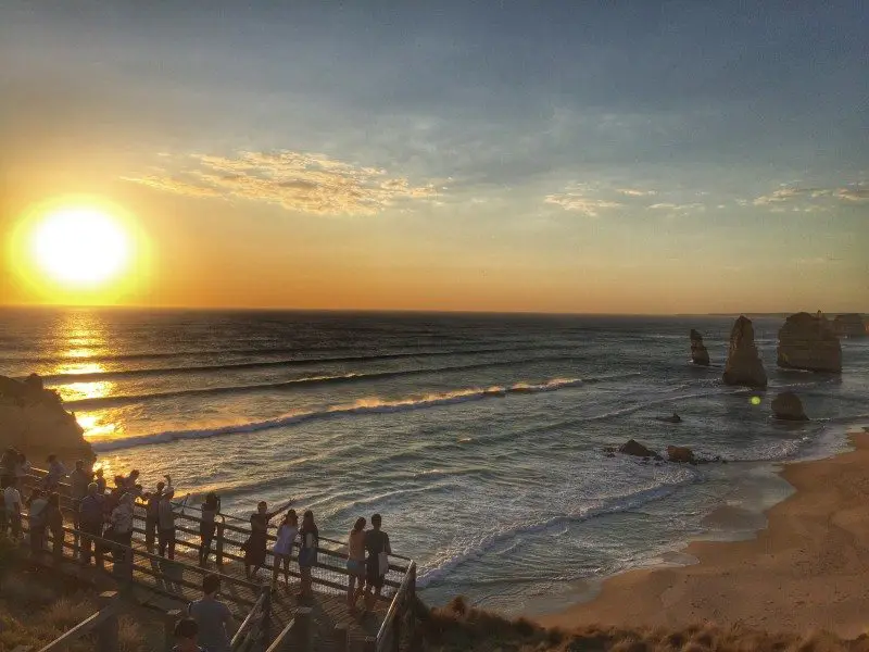 Pessoas em um calçadão assistindo ao pôr do sol sobre o oceano, perto de formações rochosas em uma praia.