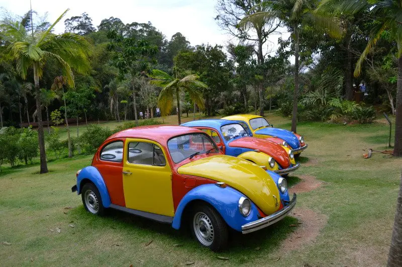 Três carros coloridos e retrô com exteriores vermelho, amarelo e azul estão estacionados em uma área gramada cercada por árvores.