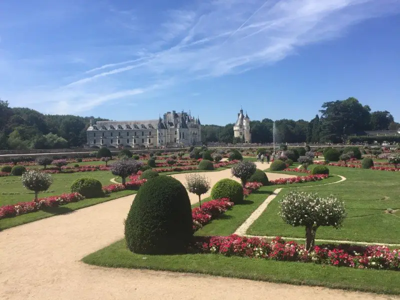 Castelo de Chenonceau com jardins bem cuidados e canteiros de flores coloridas sob um céu azul claro.