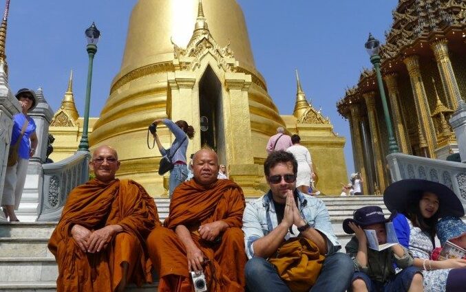 Dois monges e um homem sentados nos degraus do Grand Palace em frente a um pagode dourado. Uma mulher ajusta uma câmera ali perto. Dia claro e ensolarado.
