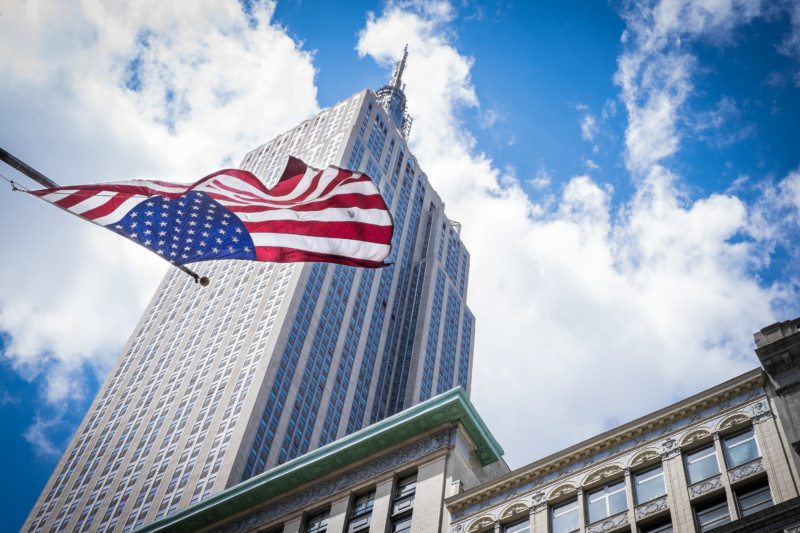 vista com ângulo de baixo para cima do empire state building. Há uma bandeira dos Estados Unidos flamulando em frente ao prédio e um céu azul com poucas nuvens ao fundo.