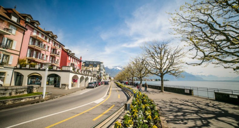 Imagem de uma rua em Montreux durante o dia com a rua do lado esquerdo da imagem e uma casas e do lado direito uma orla com algumas árvores e ao fundo um lago. Representa o que fazer em Montreux.