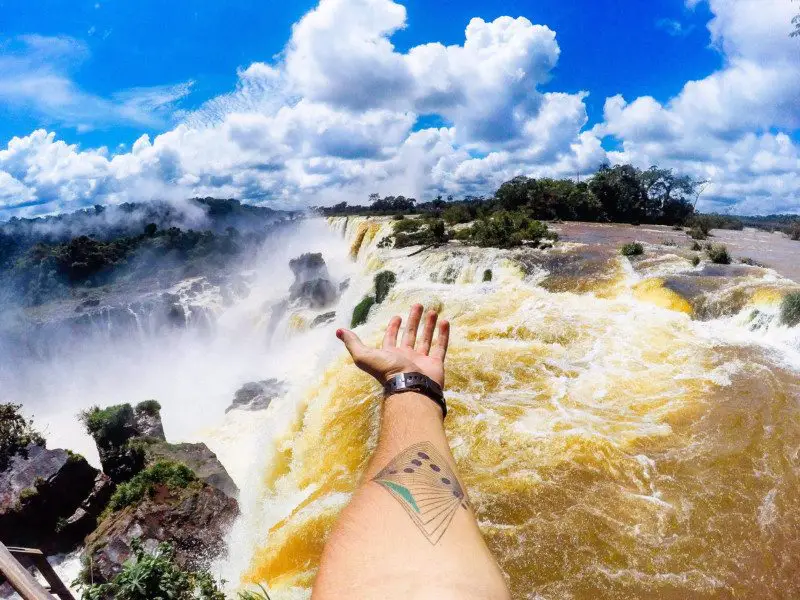 mão masculina branca estendida no centro da imagem, mostrando as cataratas de iguaçu que correm quedas abaixo com grande força. O céu está azul e conta com algumas nuvens brancas.