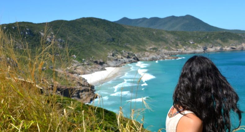 Uma pessoa com longos cabelos escuros está de frente para uma paisagem costeira em Arraial do Cabo. A praia a frente tem água azul e é cercado por areia branca e morros verdes. A foto ilustra o post de o que fazer em Arraial do Cabo.