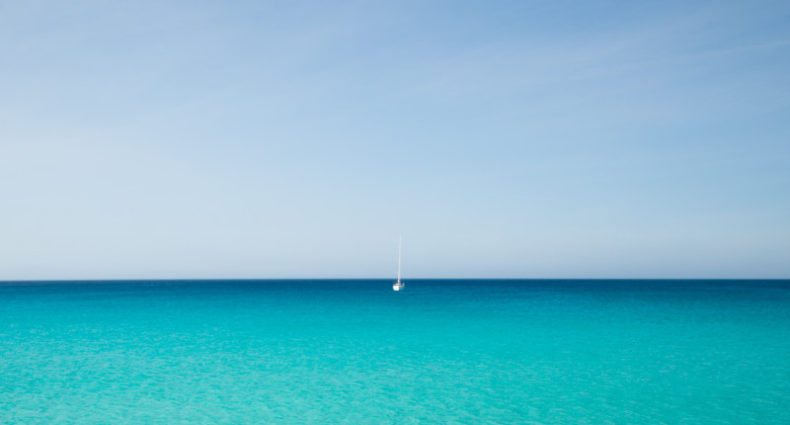 Um veleiro flutua em um mar calmo e azul-turquesa sob um céu azul claro, posicionado perto do horizonte, no centro da imagem. Essa é a praia de Ses Platgetes, uma das opções de o que fazer em Formentera.