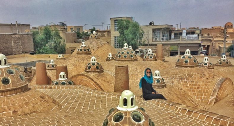 Uma pessoa sentada em um telhado de tijolos tradicional com cúpula e aberturas circulares de vidro, cercada por um céu nublado e edifícios no Sultan Amir Ahmad Bathhouse. Imagem para ilustrar post sobre o que fazer no Irã.