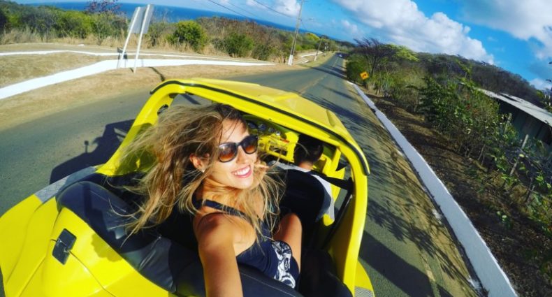 Pessoa tirando uma selfie em um conversível amarelo em uma estrada panorâmica com céu limpo e vegetação ao fundo.