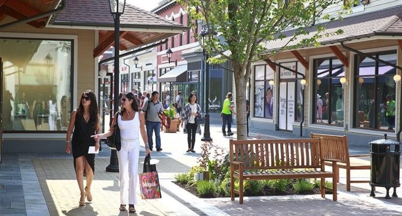 Pessoas caminhando por uma rua de compras com árvores, bancos e vitrines. Duas mulheres estão em primeiro plano, uma delas carregando uma sacola.