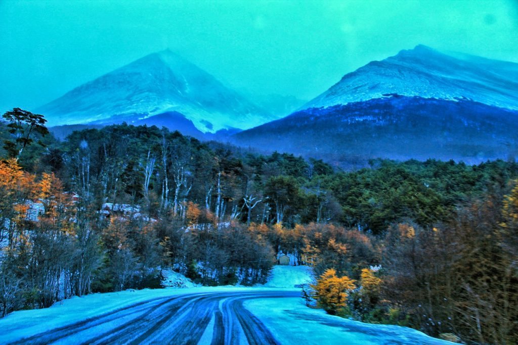 paisagem de duas montanhas nevedas com uma estrada de asfalto que leva até elas no centro da imagem, Ao redor da estrada há árvores em tons de verde e amarelo, representando o outono em ushuaia