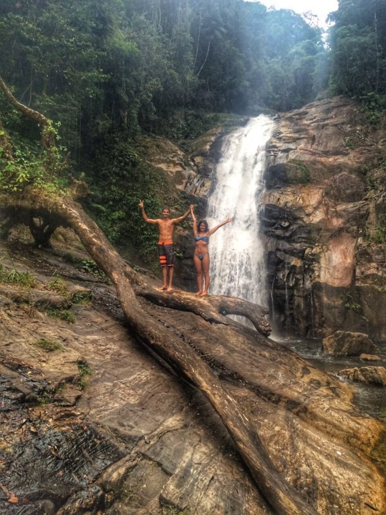 Duas pessoas em trajes de banho estão com os braços levantados sobre um grande tronco em frente a uma cachoeira cercada por vegetação exuberante. A cachoeira faz parte de um circuito acessado por uma das trilhas em SP.