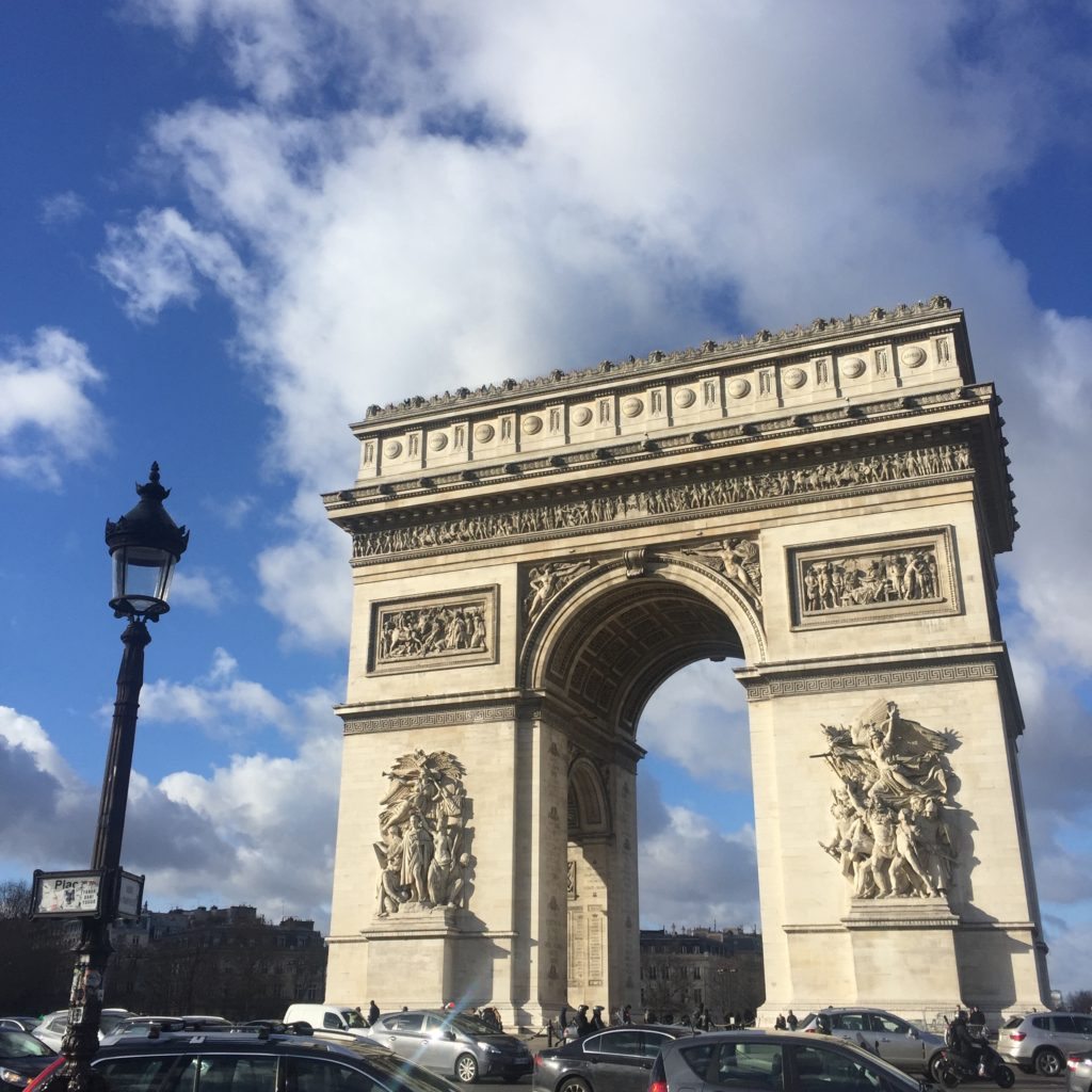 Arco do Triunfo, uma das opções de o que fazer em Paris, visto de baixo com o céu azul logo acima. Vários carros e motos circulam o ponto turístico.