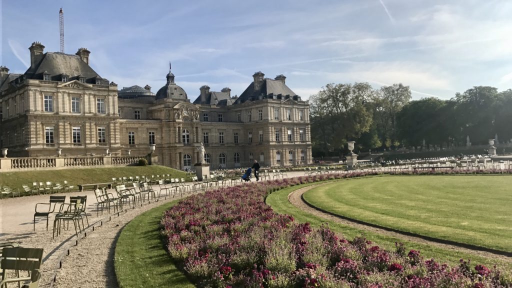 Flores coloridas ao redor de um gramado com um palácio de fundo no Jardim de Luxemburgo, uma das opções de o que fazer em Paris. Vários bancos de metal estão dispostos ao redor do gramado, e há mais árvores espalhadas pelo local. 