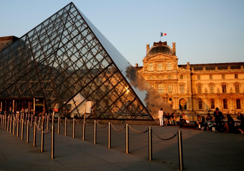 Pirâmide de vidro em frente ao Museu do Louvre, uma das opções de o que fazer em Paris. Há um caminho indicado para os viajantes, que estão sentados em bancos ao lado da instalação. 