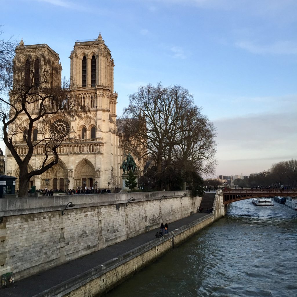 Basílica de Notre-Dame, uma das opções de o que fazer em Paris. A igreja está ao lado esquerdo da imagem, perto de algumas árvores. Ao lado direito está o rio Sena.