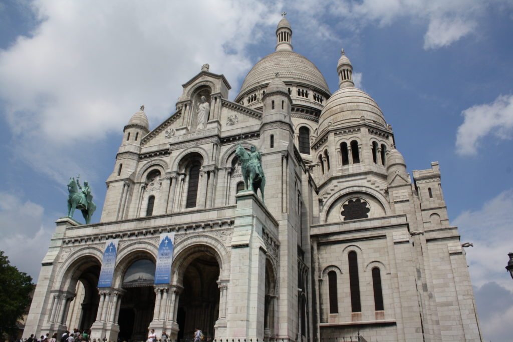 Fachada da Basílica de Sacré-Coeur, uma das opções de o que fazer em Paris. Vários domos compõem o local, que tem esculturas de cavaleiros à frente.