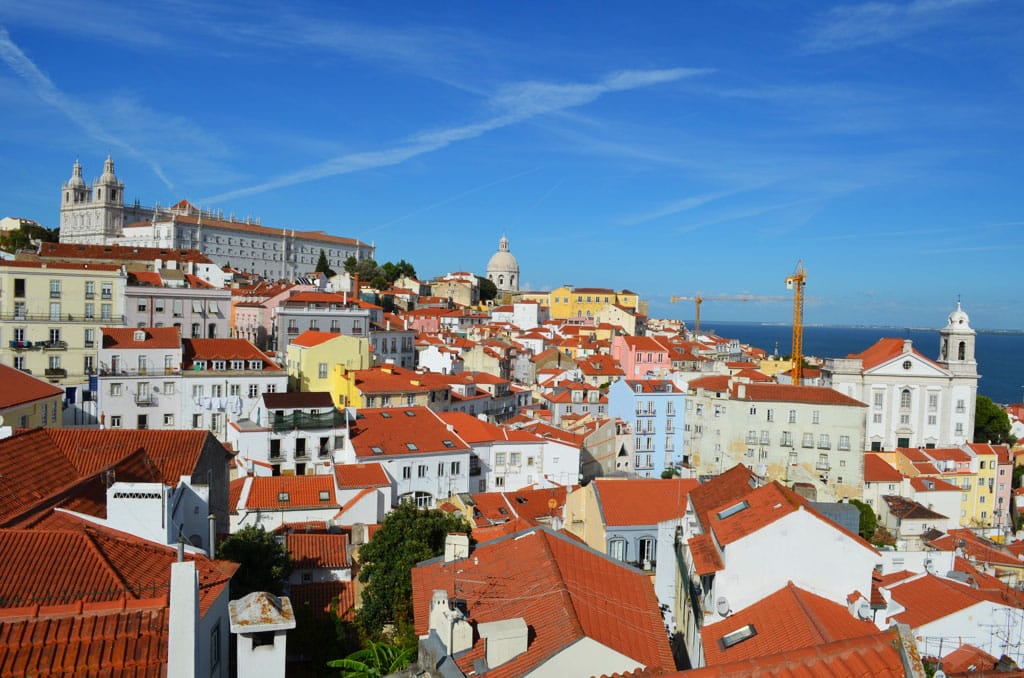 Vista de aérea do centro histórico de Lisboa, em dia de céu limpo, com casinhas de telhado laranja e mar ao fundo.