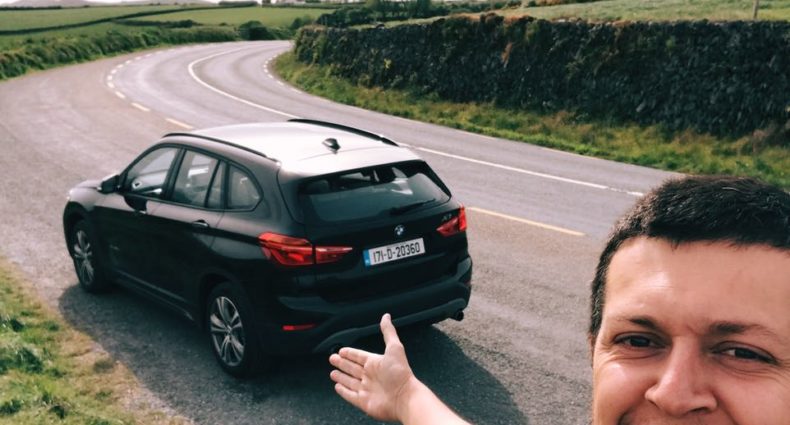 Flávio Antunes está parado na beira da estrada gesticulando para um carro preto em uma estrada sinuosa através de uma paisagem verdejante na Irlanda