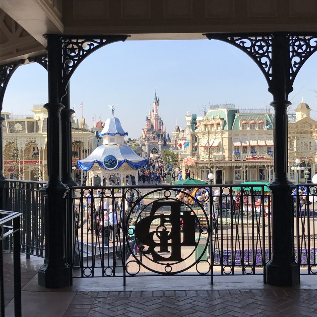 Visão da Disneyland Paris, uma das opções de o que fazer em Paris, de um local coberto. Ao fundo é possível ver o Castelo da Bela Adormecida com suas torres, e outras construções ao redor. Várias pessoas caminham ali.