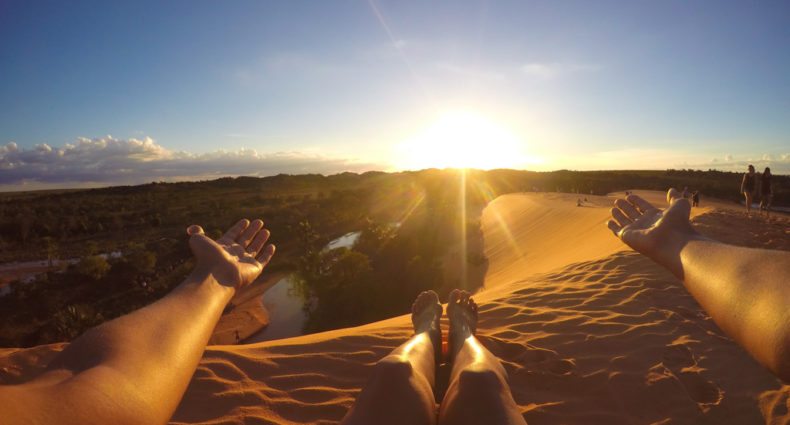 Uma pessoa sentada nas Dunas do Jalapão com os braços estendidos, de frente para o pôr do sol. Um rio e vegetação são visíveis ao fundo. Foto para a lista de o que fazer no Jalapão.