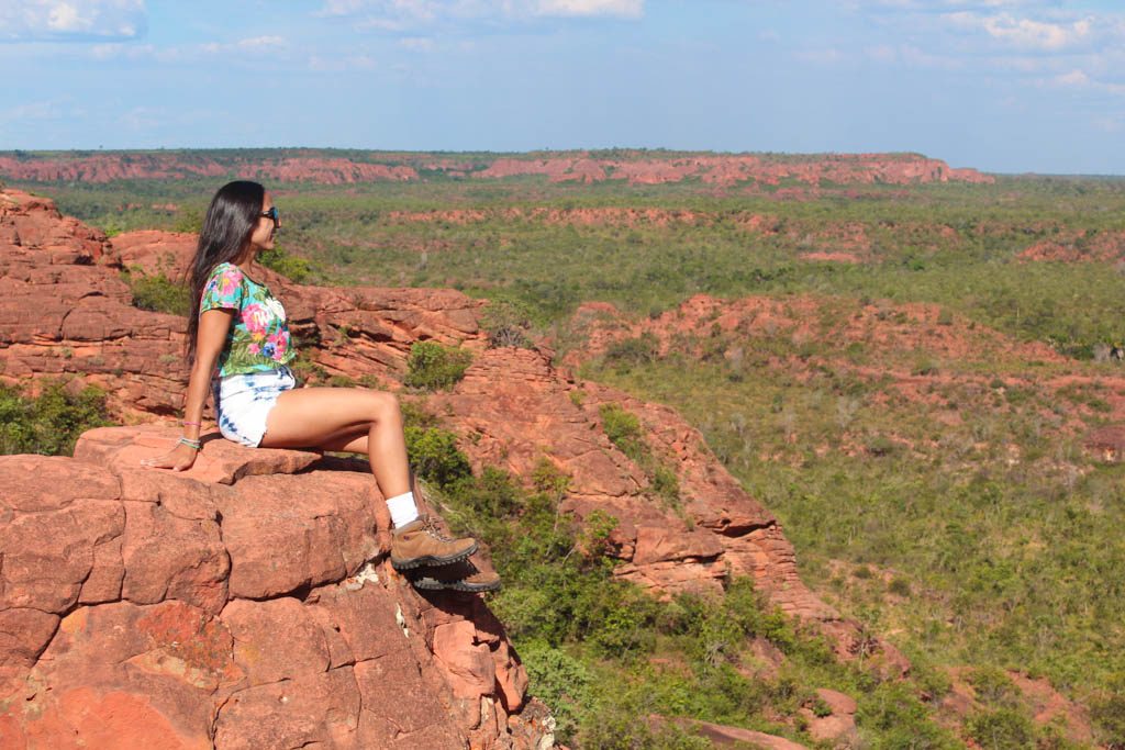 Uma mulher está sentada na beira do Morro Vermelho com vista para uma vasta paisagem de terreno verde e vermelho sob um céu parcialmente nublado. Foto para a lista de o que fazer no Jalapão.