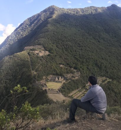 Vista do alto das ruinas de Choquiquerao. Na imagem, tem um homem sentado em uma pedra, olhando para a paisagem, que é composta por montanhas em volta de uma cidade inca no meio
