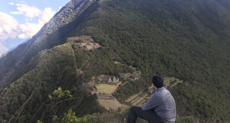 Vista do alto das ruinas de Choquiquerao. Na imagem, tem um homem sentado em uma pedra, olhando para a paisagem, que é composta por montanhas em volta de uma cidade inca no meio