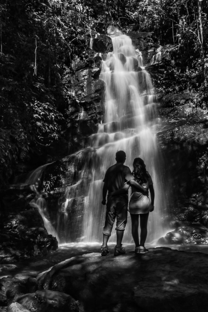 Casal de frente para a Cachoeira Véu da Noiva em Visconde de Mauá. A cachoeira é em um paredão grande com algumas árvores em volta.