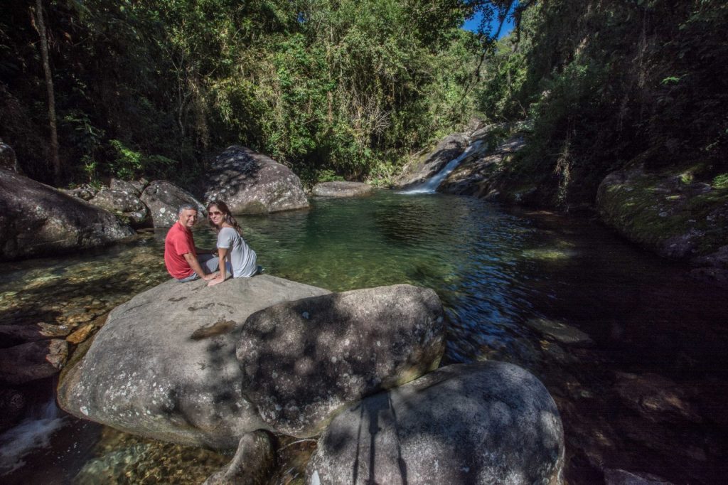 Poço do Marimbondo com várias pedras e mata fechada ao redor. Na pedra da frente do poço tem um casal sentado durante o dia. Imagem ilustrando post O que fazer em Visconde de Mauá.