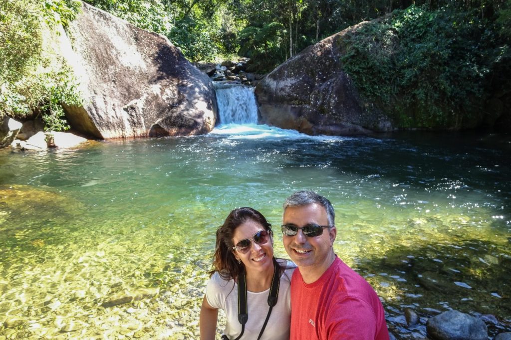 Casal tirando foto no Poção de Maromba com águas verdes azuladas, pedras grandes no fundo, uma pequena queda d'água e mata fechada ao redor. 