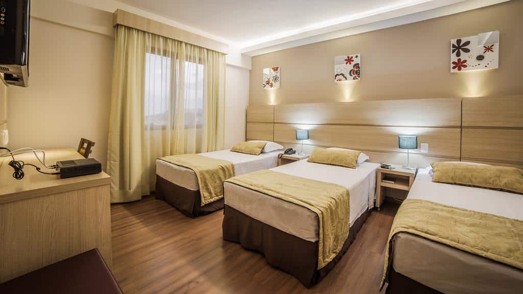 Quarto do Faro Hotel com  três camas de solteiro do lado direito em frente as camas uma cômoda de madeira com TV pendurada na parede.