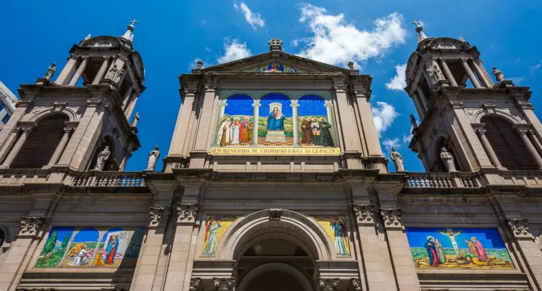 Vista frontal de uma catedral com duas torres sineiras, com mosaicos religiosos coloridos na fachada sob um céu azul.