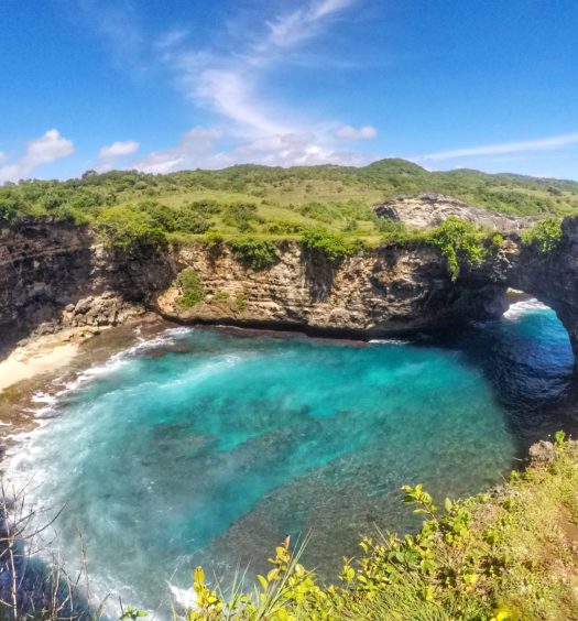 Praia de Broken Beach, em Nusa Penida. A foto mostra o arco de pedra natural sobre águas cristalinas azul-turquesa do lado direito, cercado por vegetação exuberante e penhascos rochosos sob um céu azul. No meio está o mar.