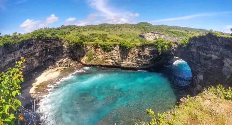 Praia de Broken Beach, em Nusa Penida. A foto mostra o arco de pedra natural sobre águas cristalinas azul-turquesa do lado direito, cercado por vegetação exuberante e penhascos rochosos sob um céu azul. No meio está o mar.