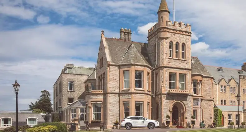 Fachada do The Culloden Estate and Spa, uma opção de onde ficar em Belfast. A imagem mostra um grande edifício de pedra com uma torre, entrada em arco e um carro branco estacionado na frente, sob um céu parcialmente nublado.