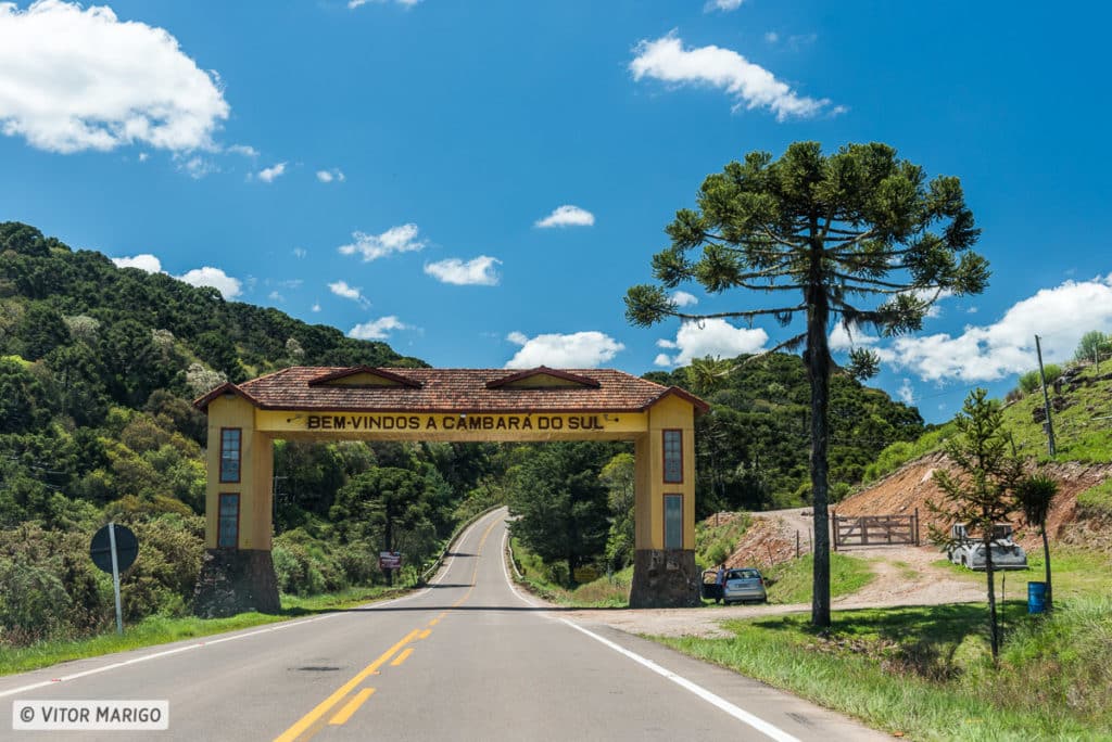 pórtico de entrada amarelo em cima da estrada asfaltada, repleto de vegetação natural, araucárias e um céu azul límpido. No portal está escrito "Bem-vindos a Cambará do Sul"