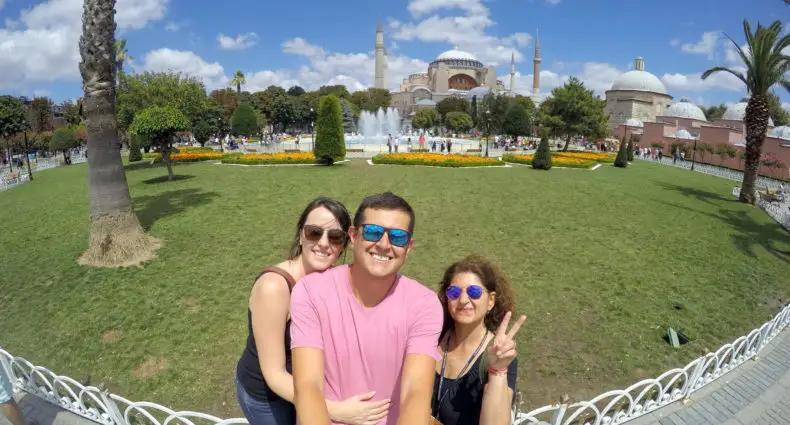 Três pessoas posam para uma selfie em frente a uma fonte e edifício histórico. O céu está claro com nuvens dispersas. A foto ilustra o post sobre guia em Istambul.