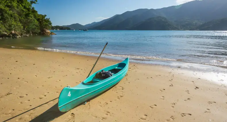 Um caiaque cor turquesa com um remo repousa em uma praia de areia com águas calmas no Saco do Mamanguá, com colinas verdes e um céu azul ao fundo.
