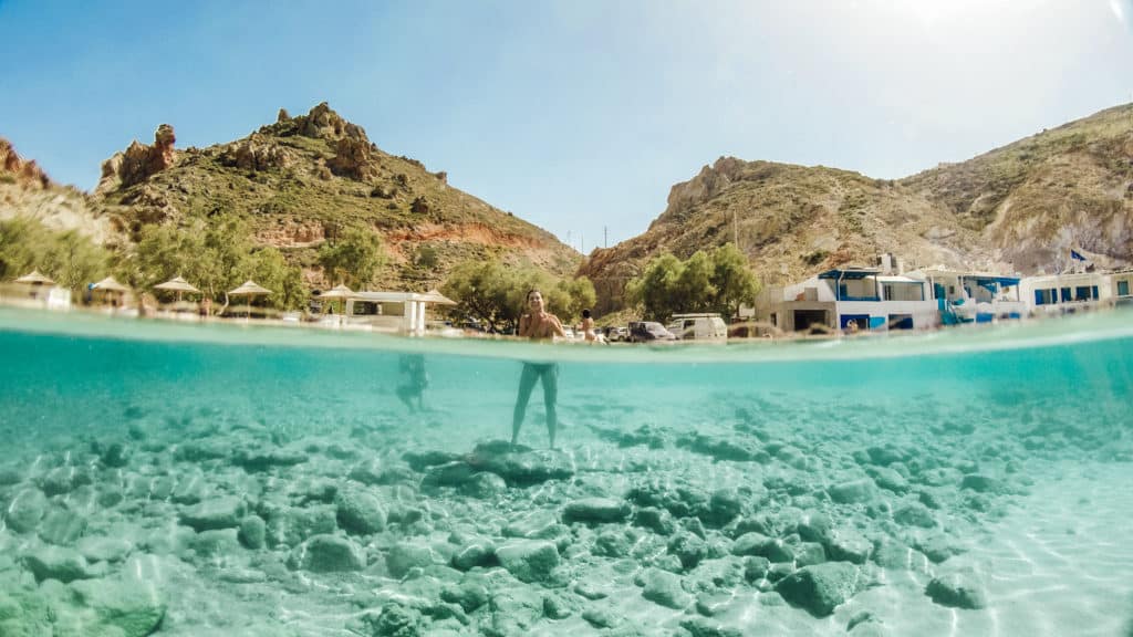 Uma imagem de visão dividida de um terreno rochoso subaquático e uma pessoa vadeando, com colinas, edifícios e árvores ao fundo sob um céu limpo.