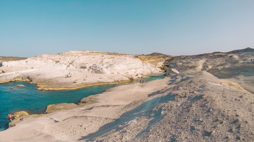 Paisagem costeira rochosa com penhascos brancos, águas azul-turquesa e algumas pessoas espalhadas pela costa nadando. Essa é uma das dicas de o que fazer na Ilha de Milos.