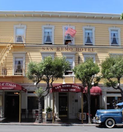 O San Remo Hotel amarelo com uma bandeira americana, mesas ao ar livre e um carro antigo estacionado na frente.