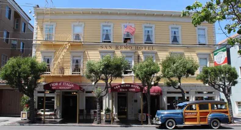 O San Remo Hotel amarelo com uma bandeira americana, mesas ao ar livre e um carro antigo estacionado na frente.