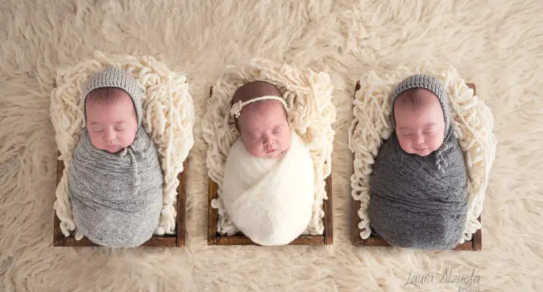 Três bebês enfaixados dormindo em caixas de madeira sobre um tapete peludo bege. Os bebês usam roupas e chapéus cinza e branco. A imagem ilustra o post sobre enxoval de bebê nos EUA.