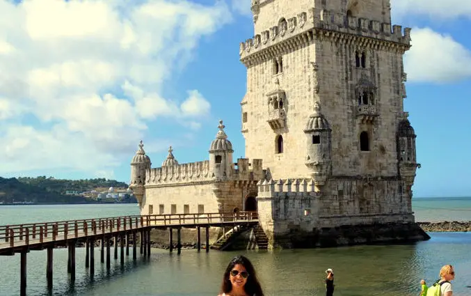 Uma pessoa sorri em frente à Torre de Belém, em Lisboa, Portugal, com um céu azul brilhante e algumas nuvens acima, para ilustrar o post de pontos turísticos de Portugal.