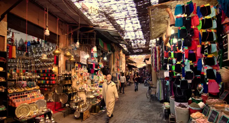 Um homem caminha pelas ruas da Medina de Marrakech movimentado com barracas exibindo tecidos coloridos, luminárias e diversos produtos sob um teto coberto. Imagem para ilustrar post sobre o que fazer no Marrocos.