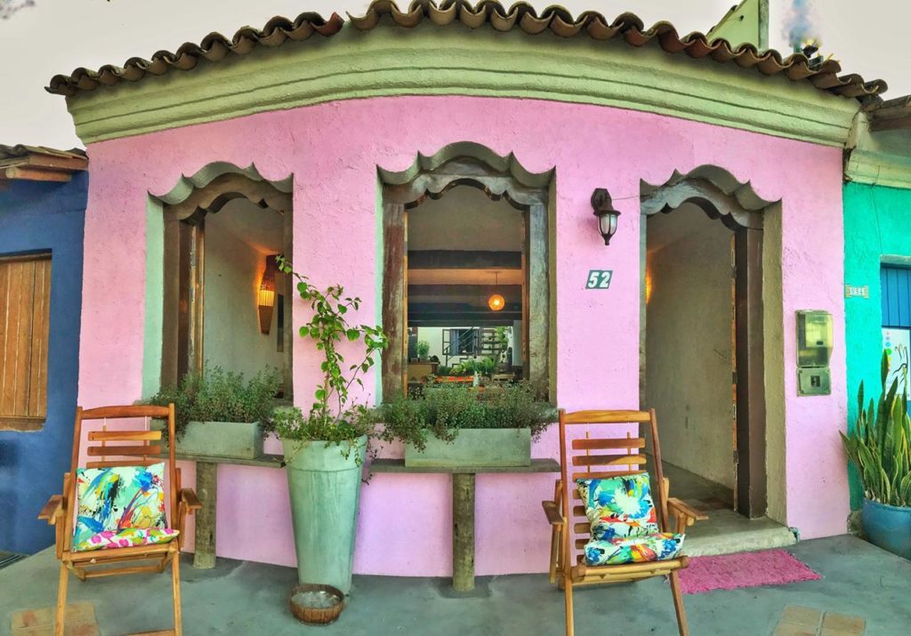 Fachada da Pousada Anacã, uma das recomendações de pousadas em Arraial D'Ajuda. A construção típica dessa região é cor de rosa e tem algumas plantas na frente. Duas cadeiras estão à frente da fachada.