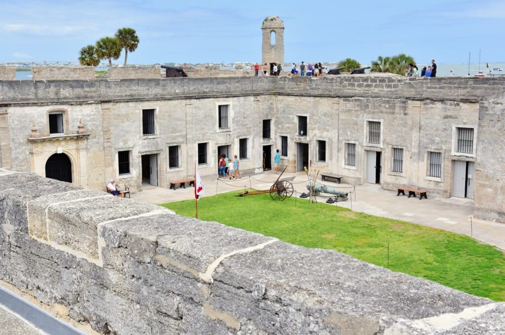 Pátio de um antigo forte de pedra com visitantes, grama verde, exibição de canhões históricos e muros de pedra ao redor sob um céu azul, uma das opções de roteiro para Flórida.