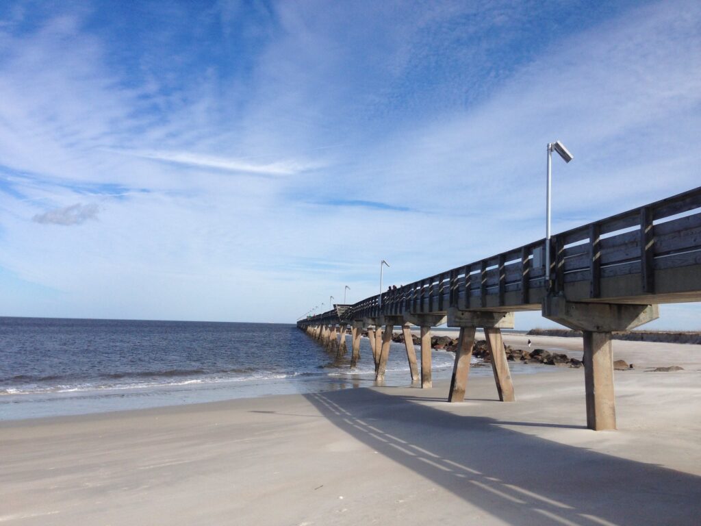Um longo píer de madeira se estende sobre um mar calmo com uma praia de areia e um céu azul parcialmente nublado ao fundo em Fort Clinch State Park, Amelia Island, uma das opções de roteiro para Flórida